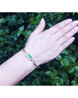 Hand wearing a bracelet with a natural emerald gemstone against a green leafy background