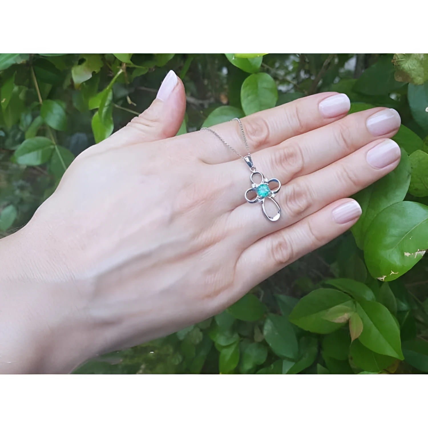 14K white gold cross pendant with a vibrant blue-green opal center, held by a hand against green leaves.