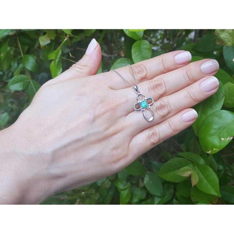 14K white gold cross pendant with a vibrant blue-green opal center, held by a hand against green leaves.