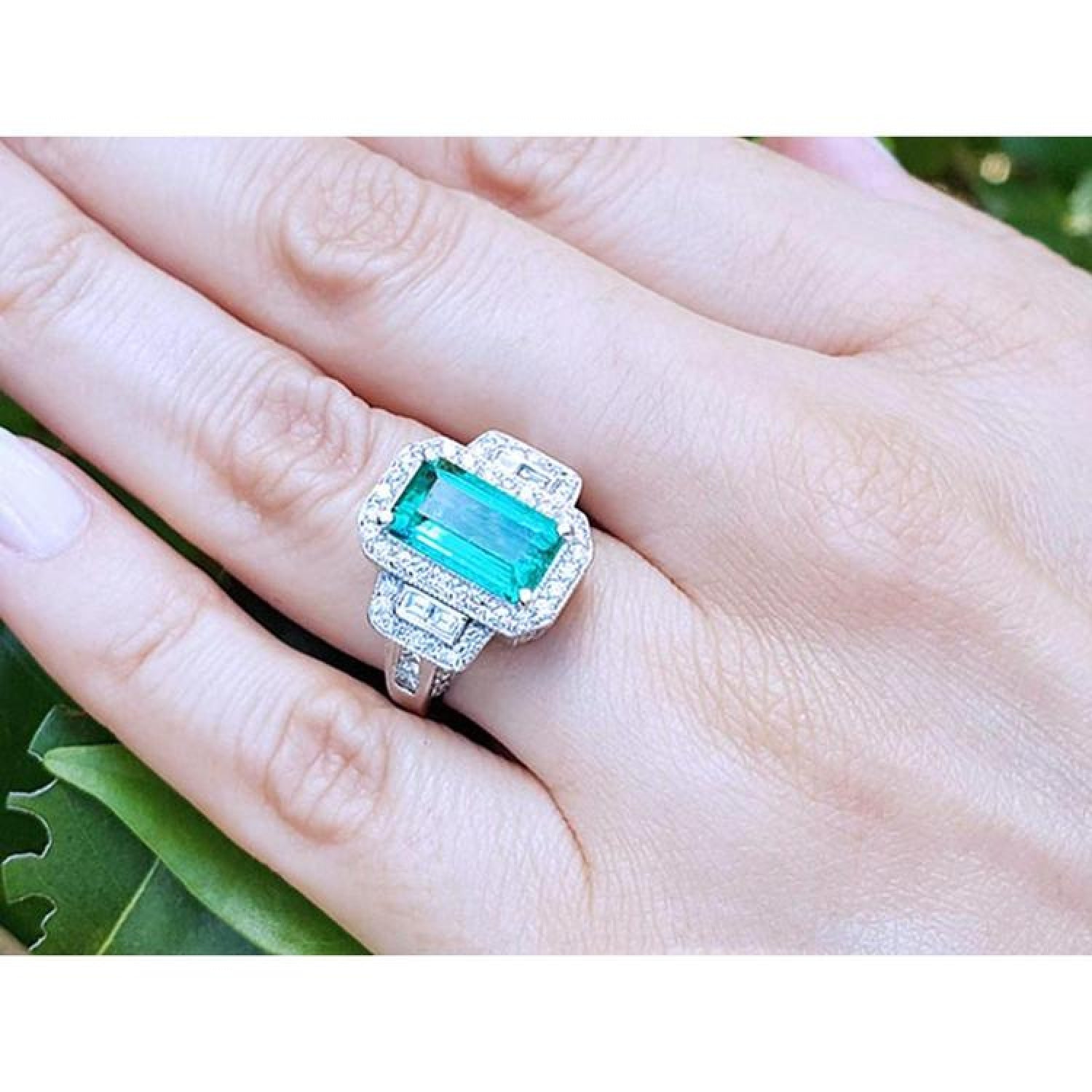 Woman's hand wearing an 18K white gold ring with a vibrant green emerald, halo, and baguette diamonds.
