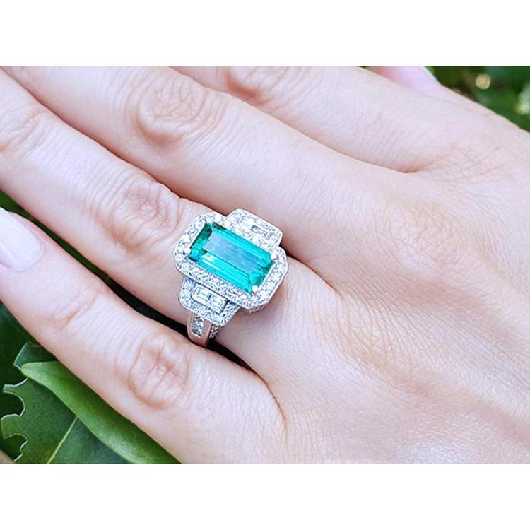 Woman's hand wearing an 18K white gold ring with a vibrant green emerald, halo, and baguette diamonds.