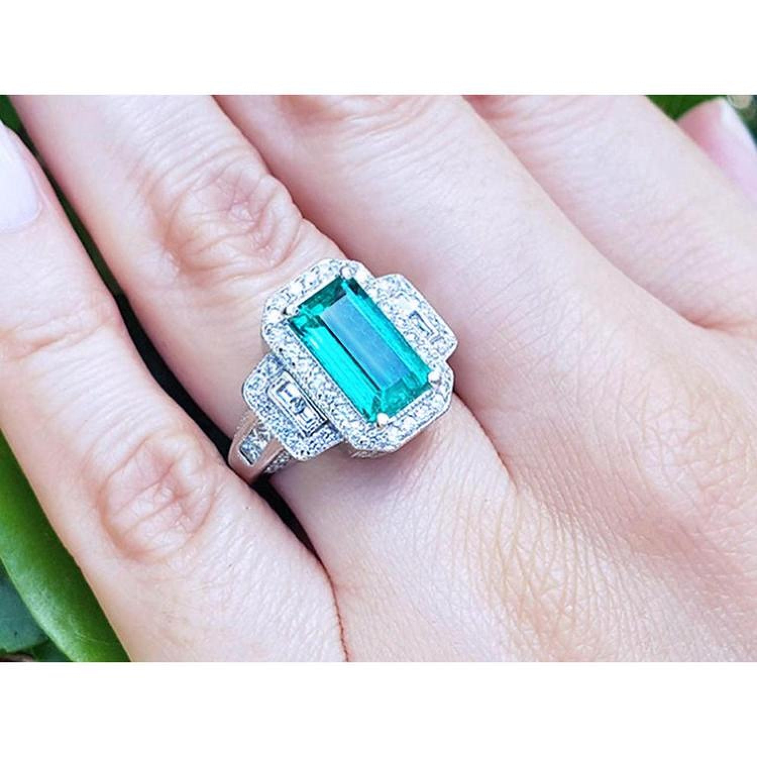 18K white gold ring with a vibrant green emerald, halo diamonds, and baguette side stones on a hand.