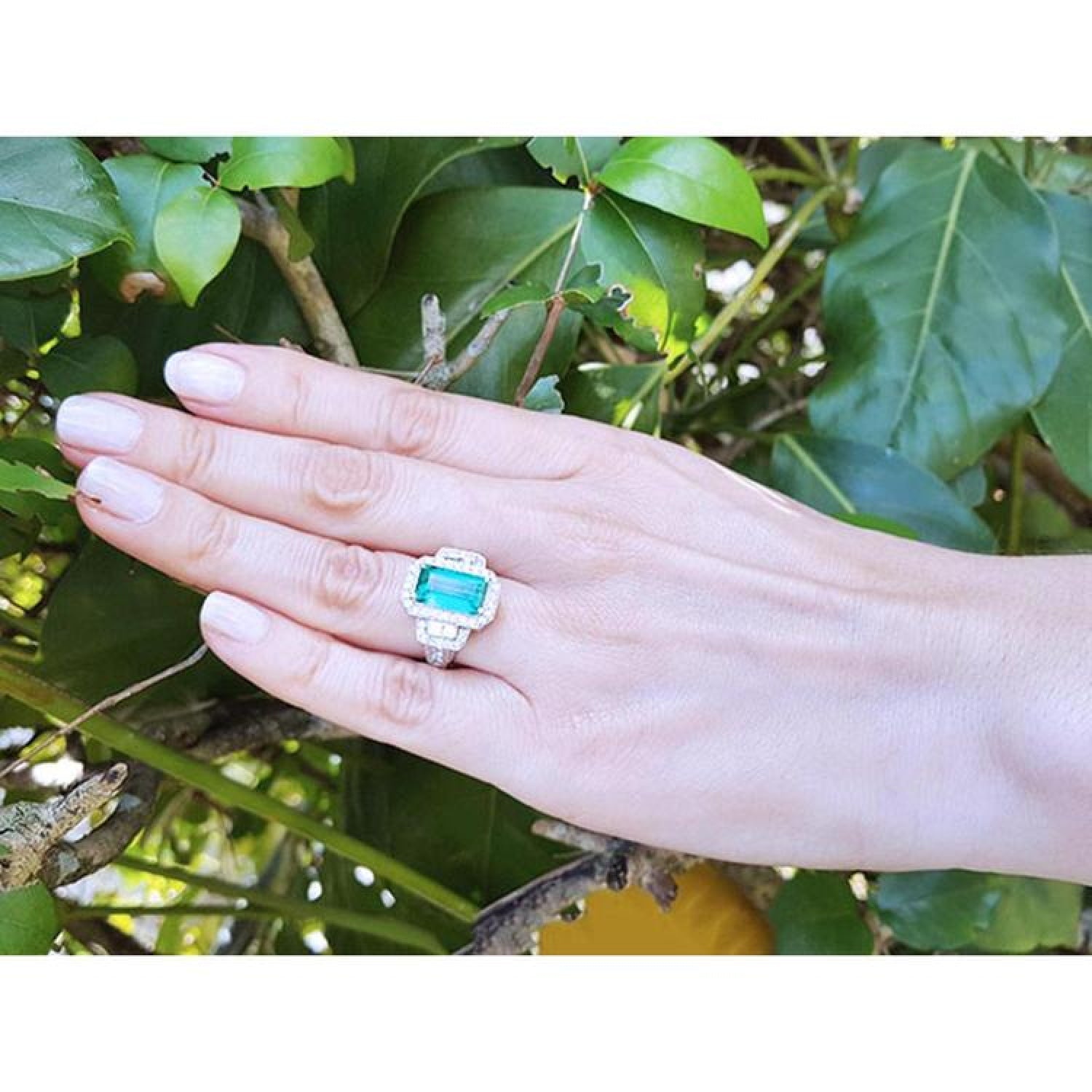 Woman's hand wearing an 18K white gold ring with a vibrant green emerald, halo diamonds, and baguette side stones.