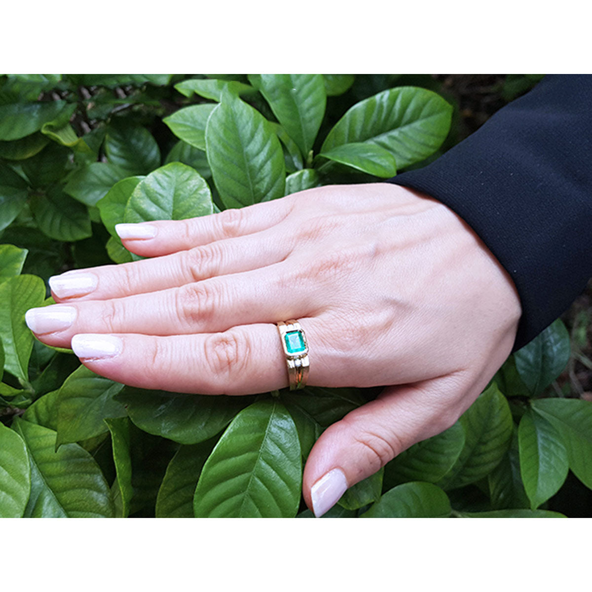 Hand wearing a gold ring for men with a natural emerald against a green leafy background.