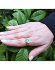 Hand wearing a gold ring for men with a natural emerald against a green leafy background.