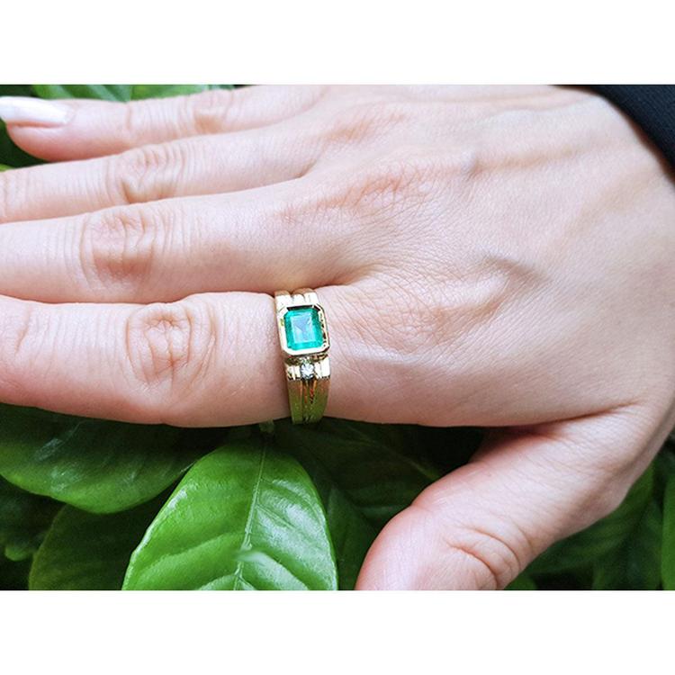 A yellow gold men's emerald ring on a hand against a leafy background.