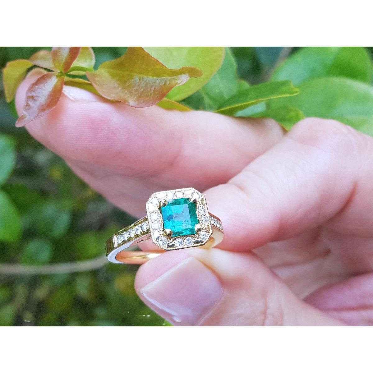 Gold ring with a square emerald and diamonds held by a hand against a green leafy background.