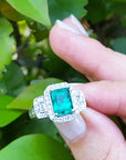 Hand holding an emerald with a halo diamond ring against a leafy background.