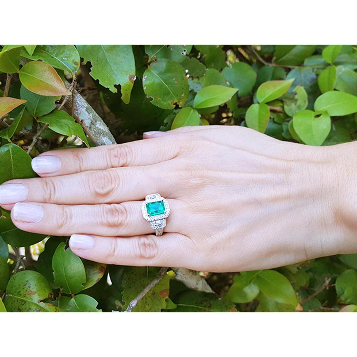 Hand wearing a natural emerald engagement ring white gold against a green leafy background