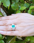 Hand wearing a natural emerald engagement ring white gold against a green leafy background