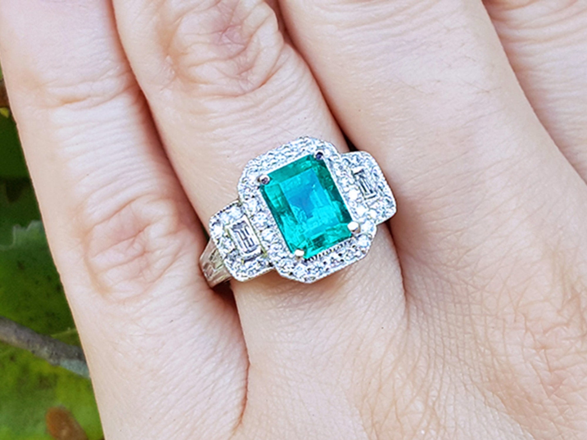 A natural emerald ring with diamond accents on a finger against a natural background.