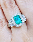 A natural emerald ring with diamond accents on a finger against a natural background.