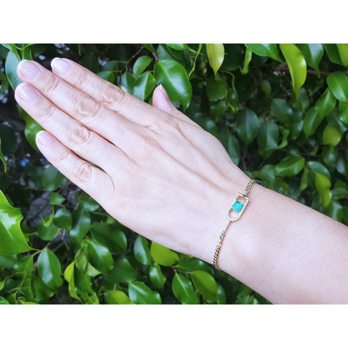 Hand wearing a gold bracelet with a emerald stone against a green leafy background.