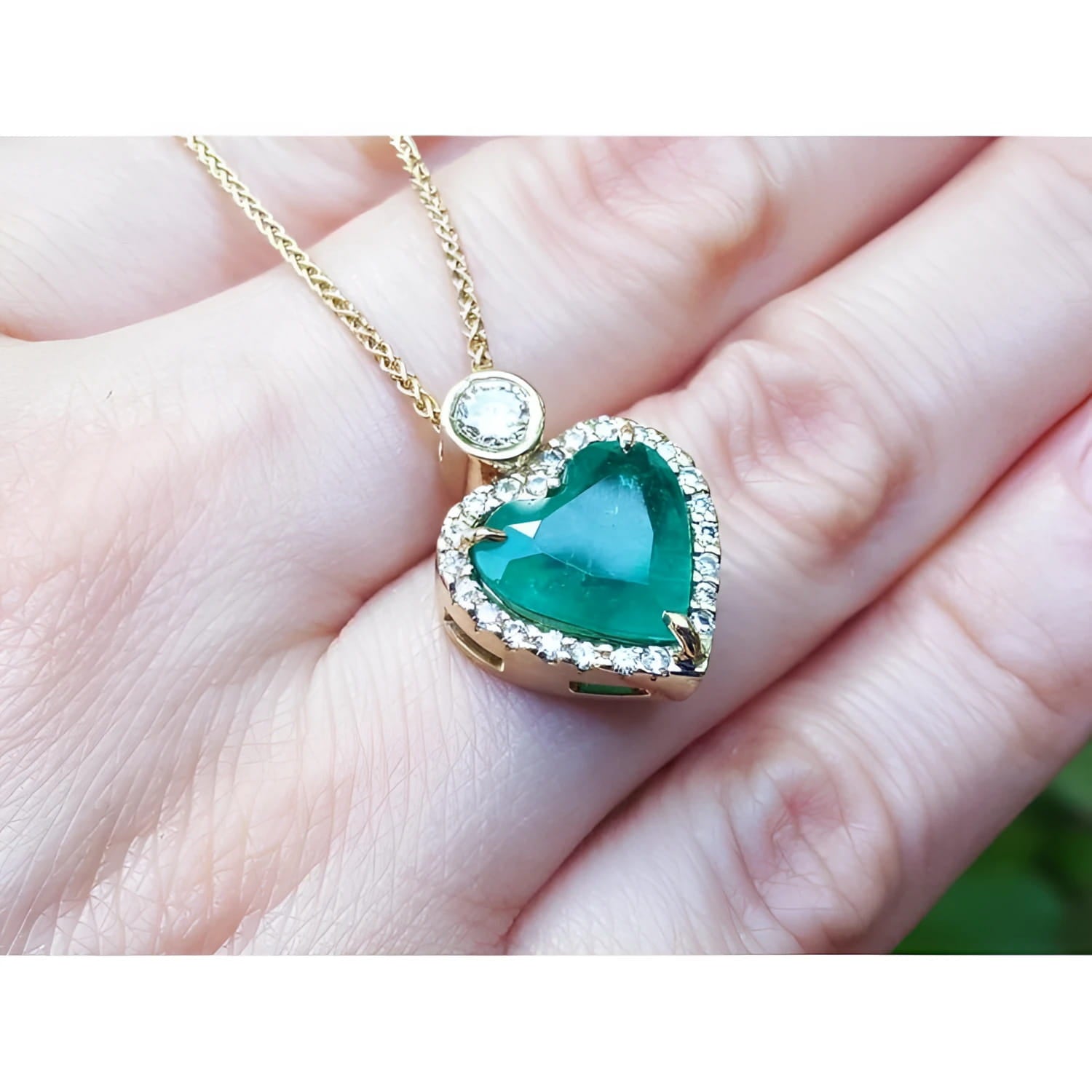 Heart-shaped emerald pendant with diamond halo on an 18k gold chain, held against a hand.