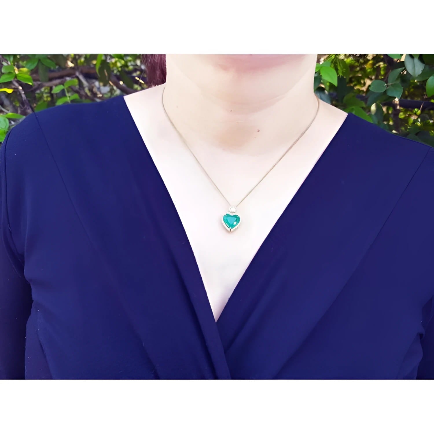 Heart-shaped emerald and diamond halo pendant in 18k gold, worn on a woman's neck.