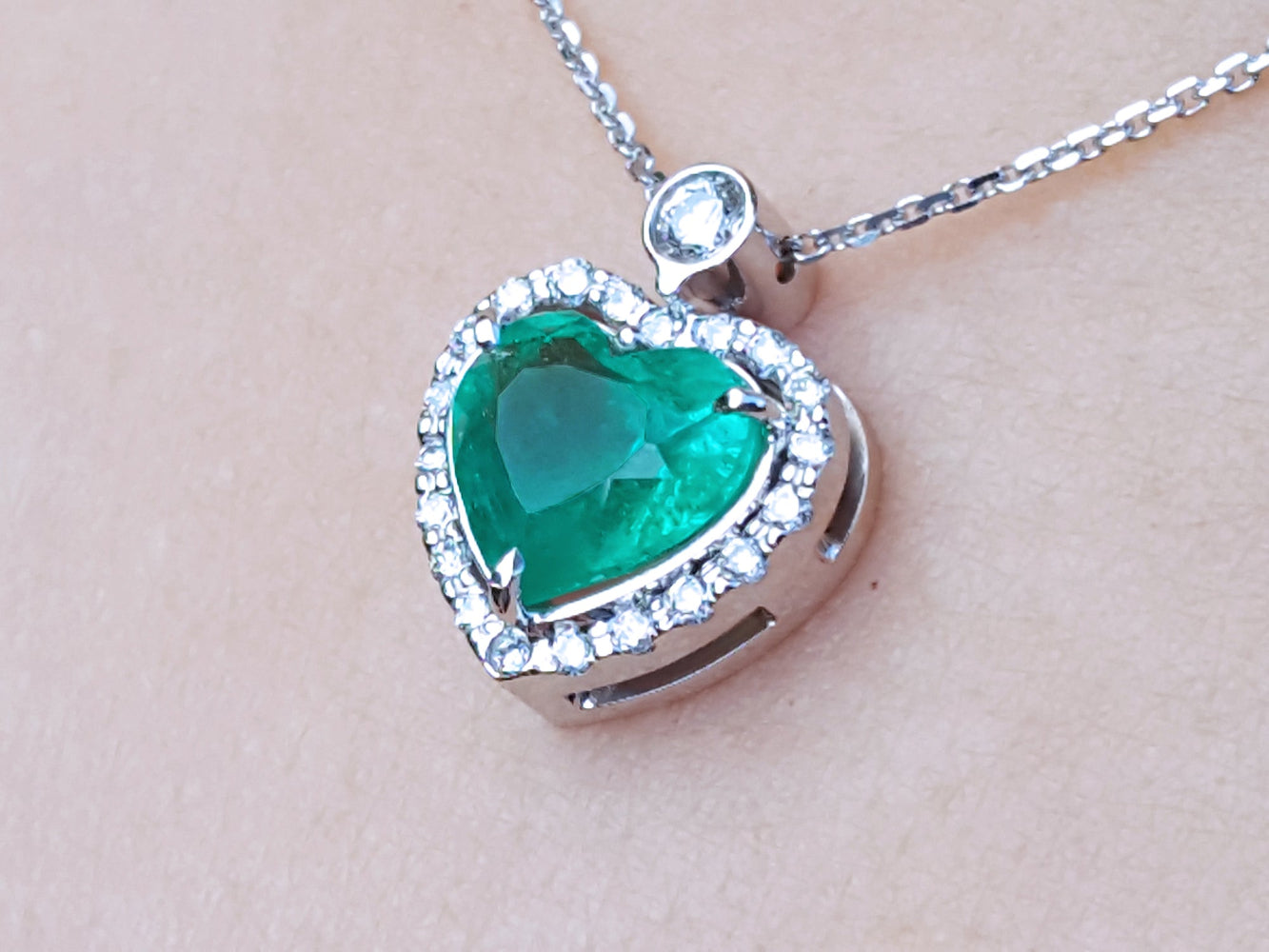 Heart-shaped emerald pendant with diamond accents on a beige skin background.