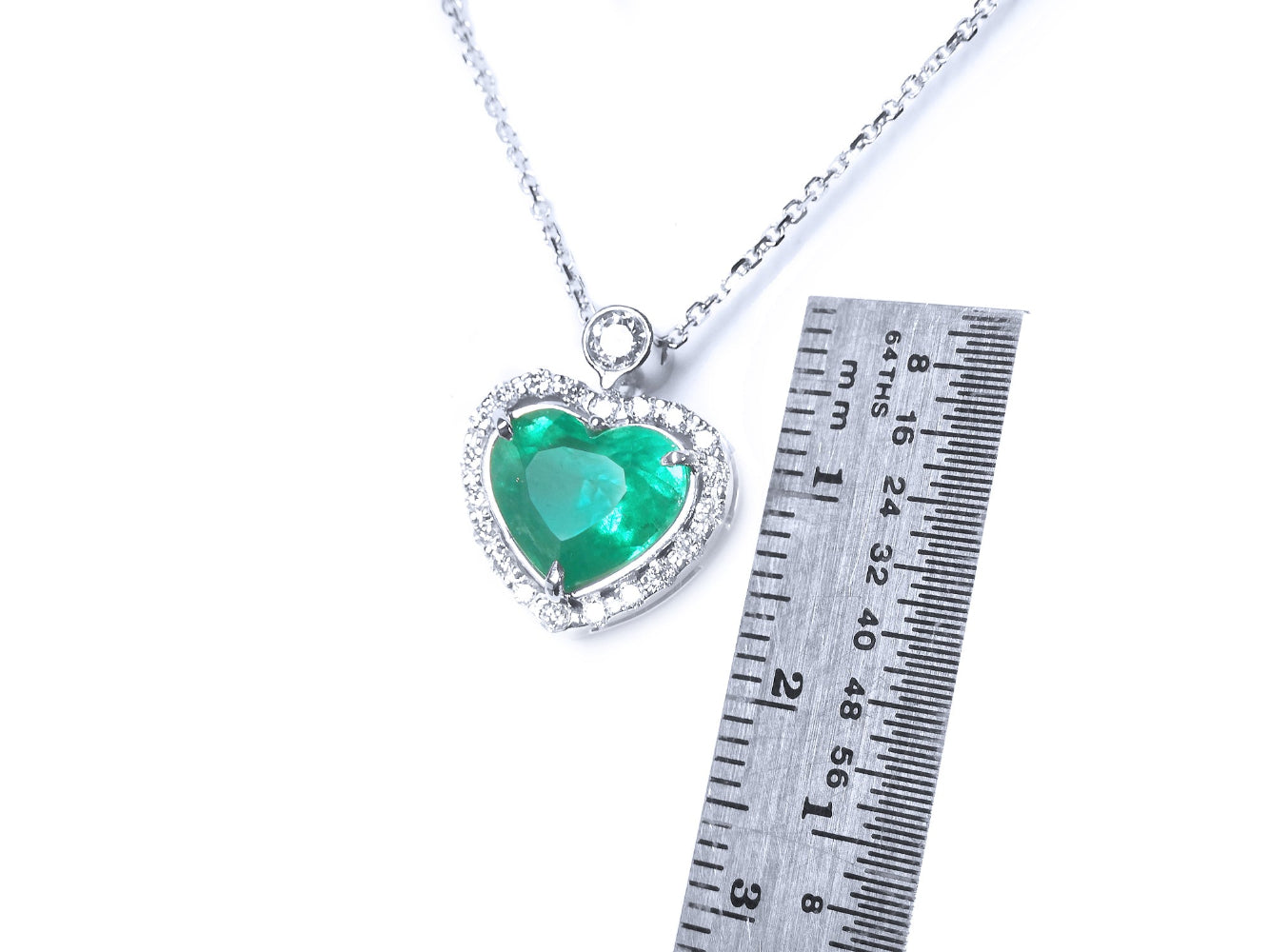 Heart-shaped emerald pendant on a chain with a ruler for scale on a white background.