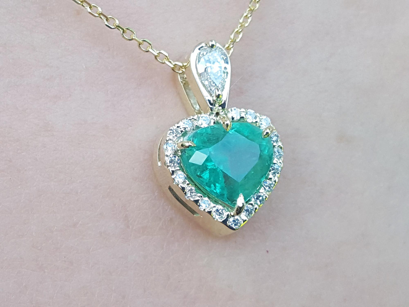 Heart-shaped emerald pendant with diamond accents on a gold chain against a light pink background