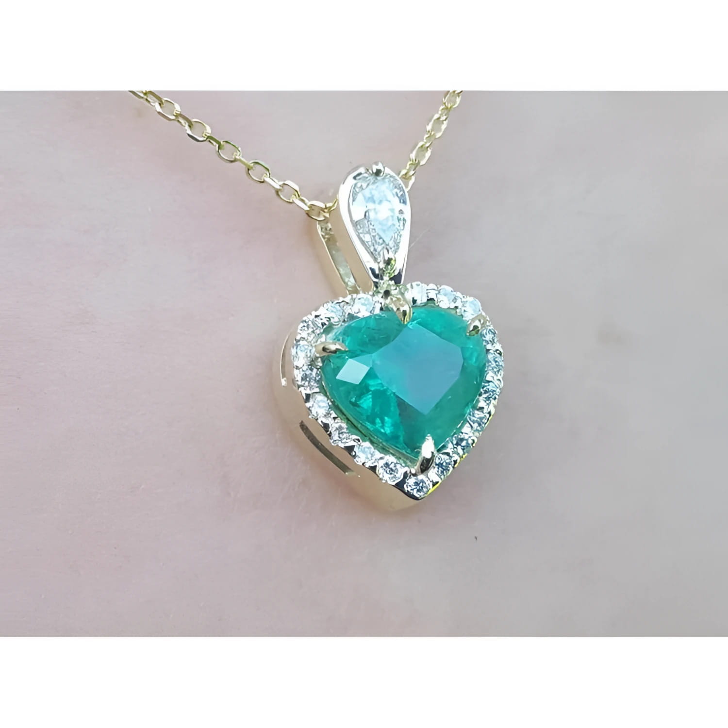 Heart-shaped emerald pendant with diamond accents on a gold chain against a light pink background