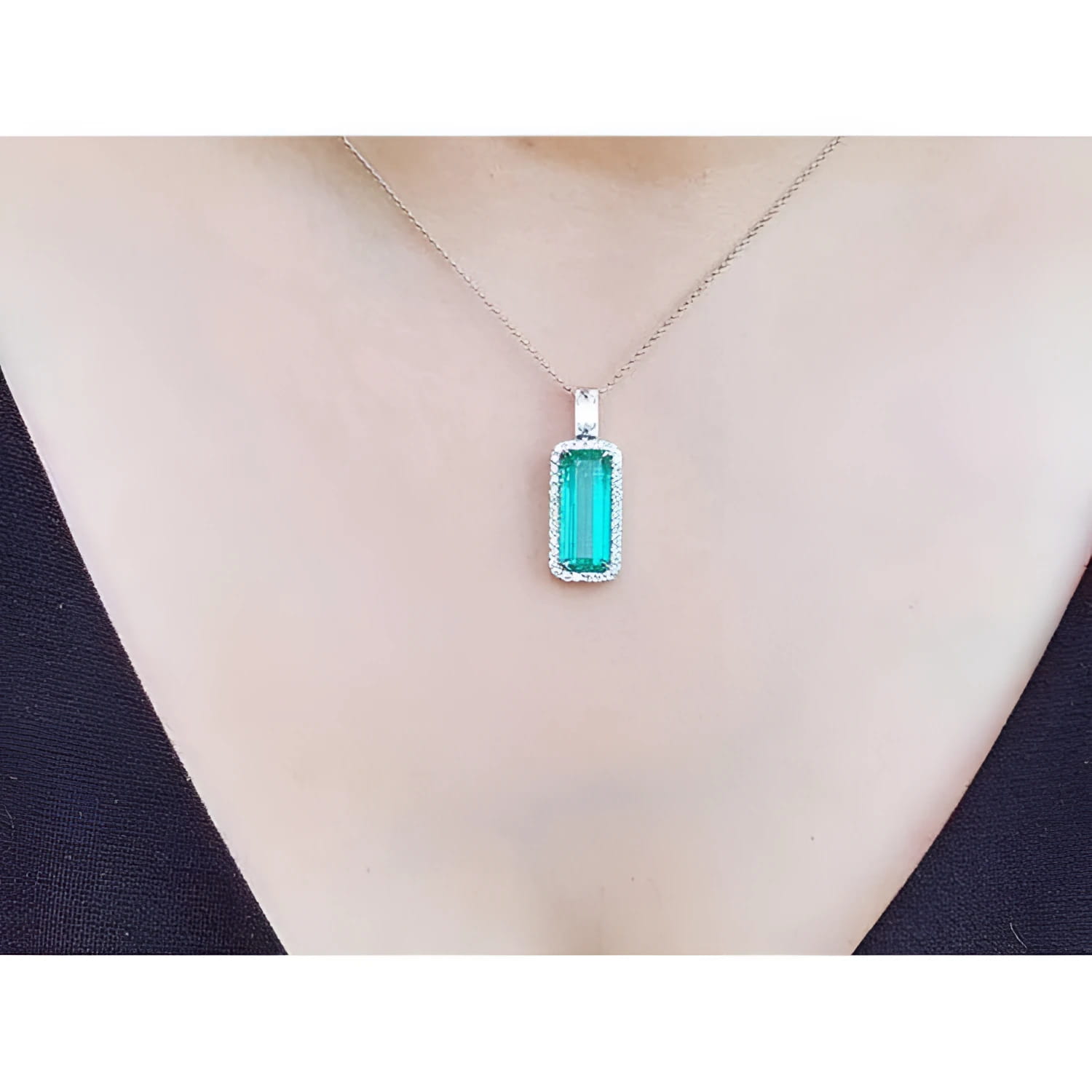 Elongated natural emerald pendant with halo diamonds on an 18K white gold chain, worn on a person.