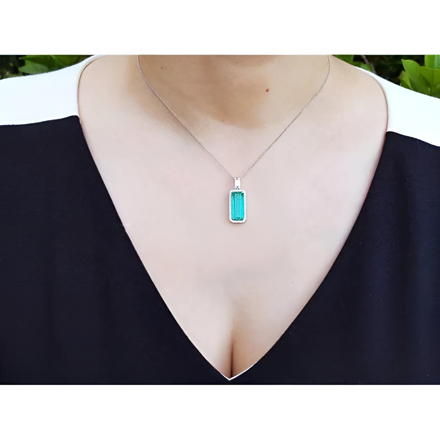 Elongated natural emerald pendant with halo diamonds on an 18K white gold chain, worn on a woman's neck.