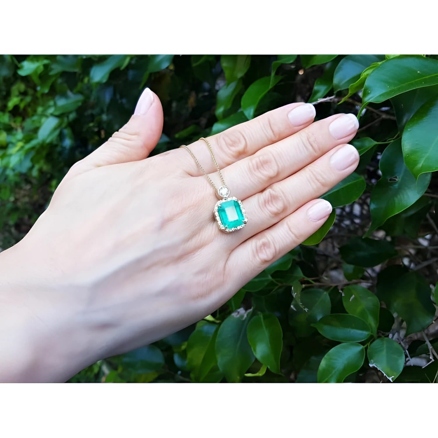18K yellow gold pendant necklace with a rectangular emerald and diamond halo, held by a hand.
