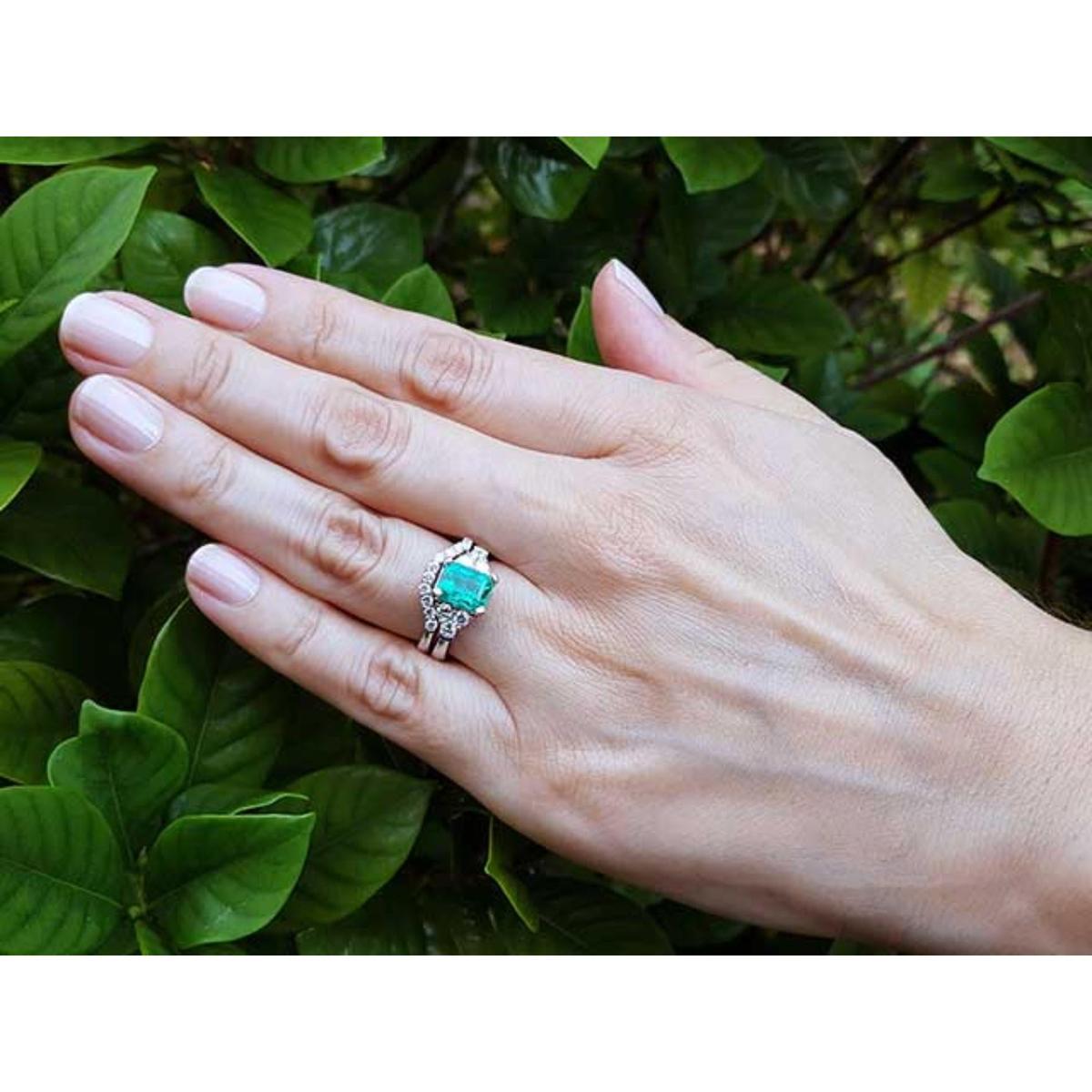 Hand wearing a natural emerald ring and matching band with green leaves in the background.