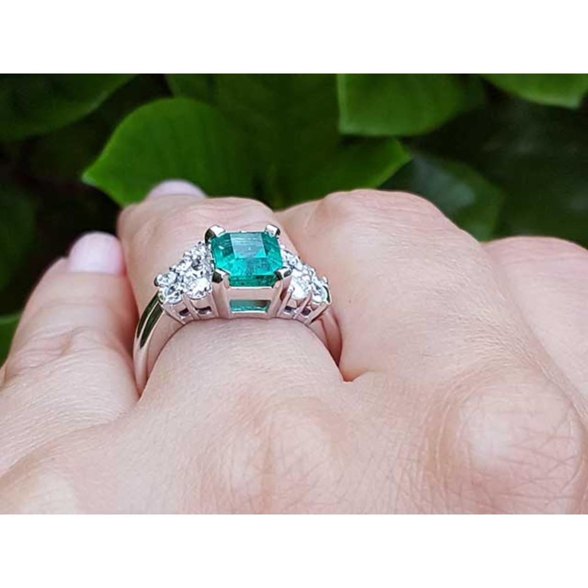 Emerald ring with diamonds and the matching band on a finger against a green leafy background.