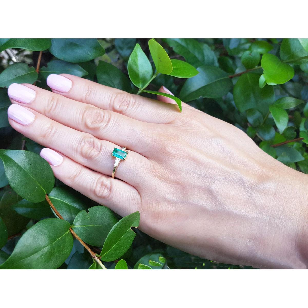 Hand wearing a ring with a natural colombian emerald against a green leafy background.