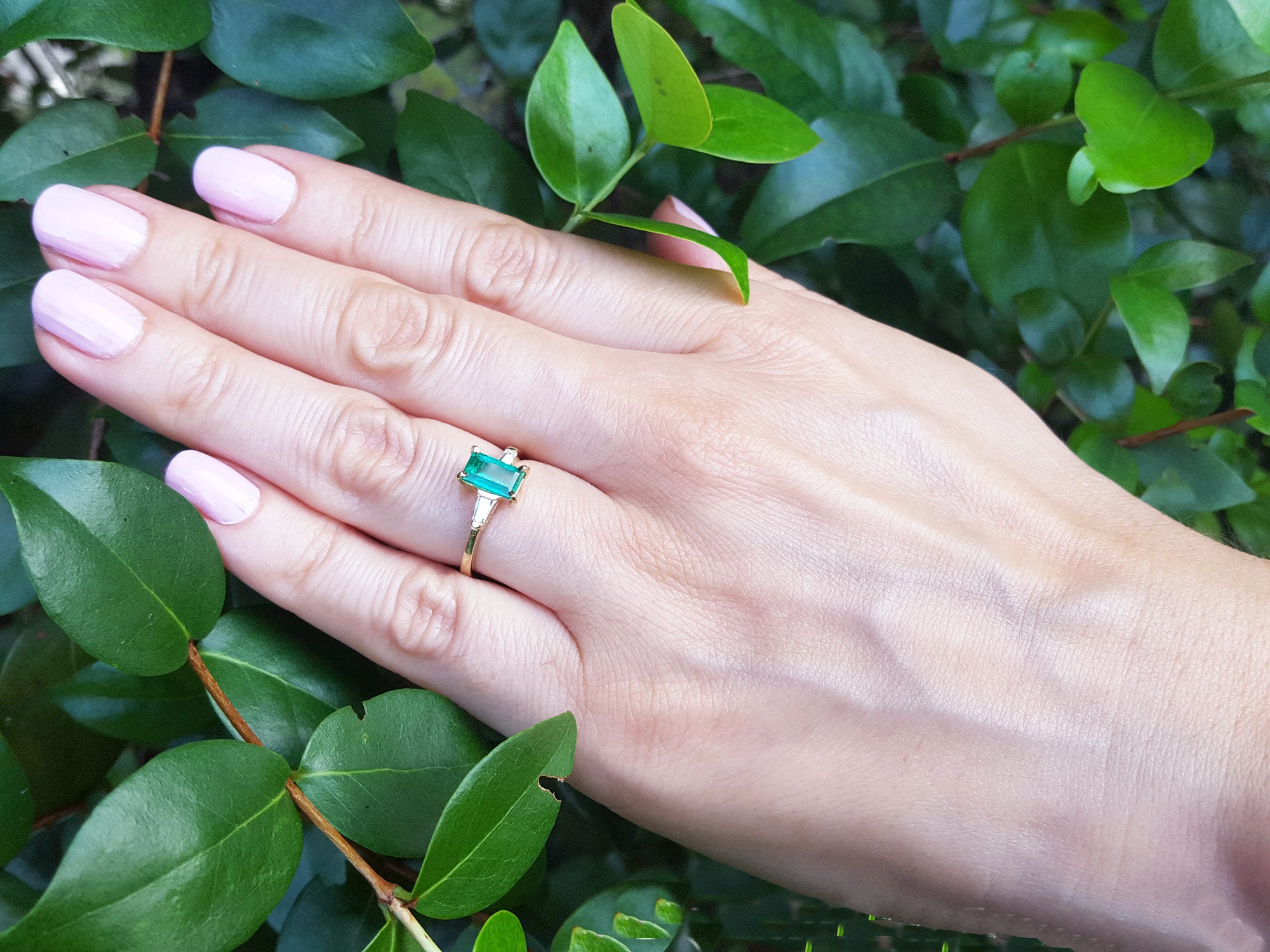 Hand wearing a ring with a natural colombian emerald against a green leafy background.