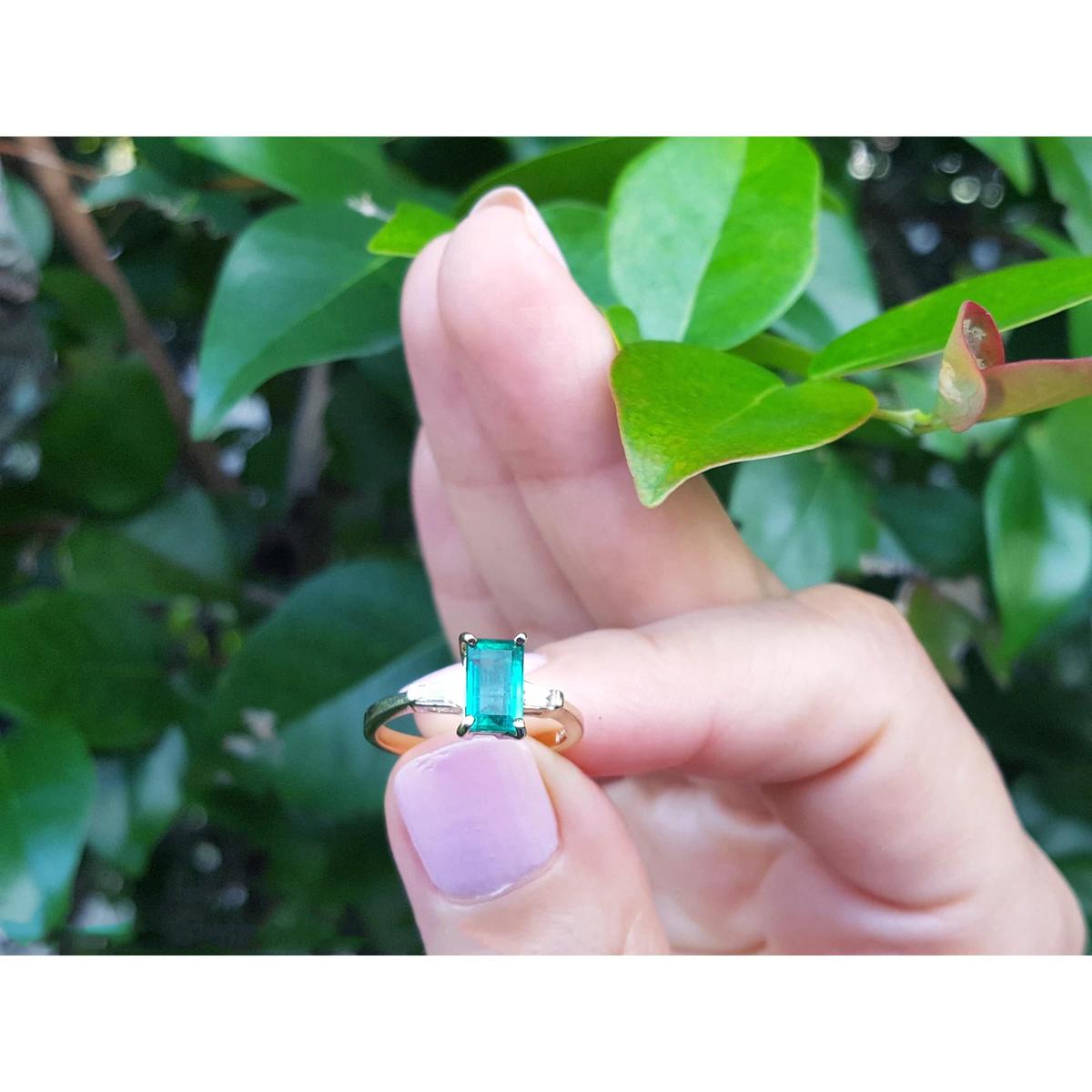 Hand holding a Colombian emerald ring with a green gemstone against a leafy background.