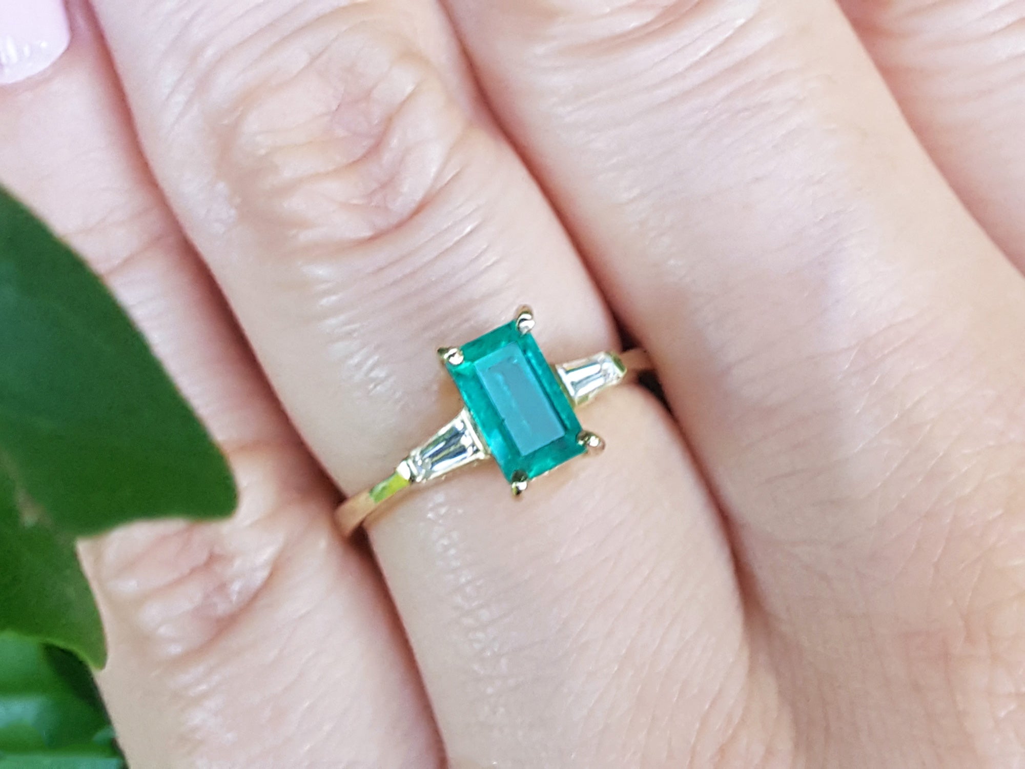 Gold ring with a natural emerald on a finger, with a blurred green leaf in the background.