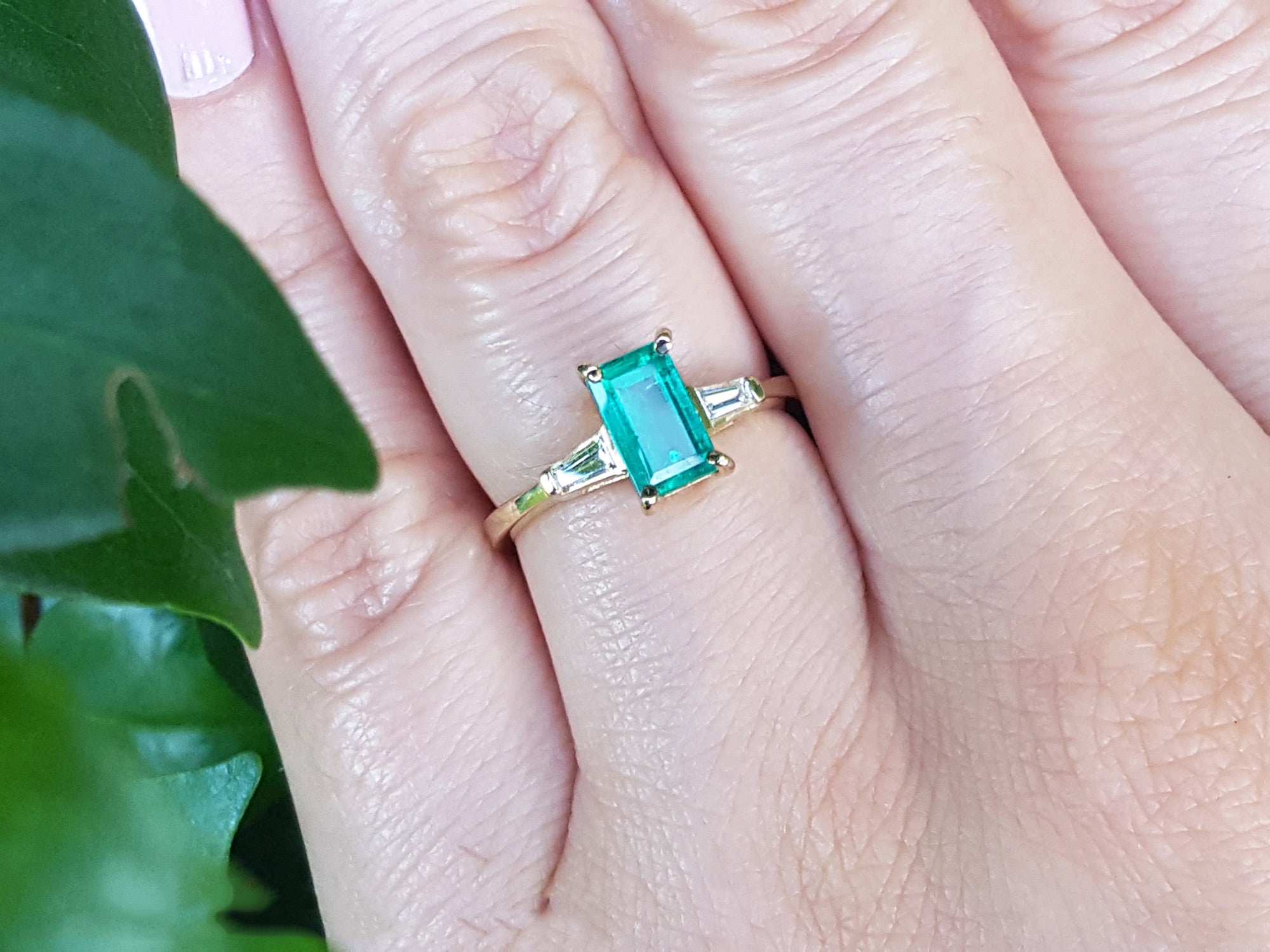 Gold ring with a real emerald on a person&#39;s finger, with a blurred green leaf in the background.
