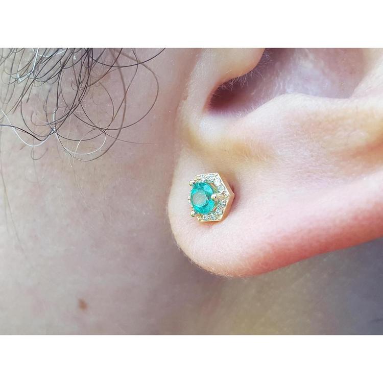 An earlobe waring an emerald and halo diamond stud earring in 14k yellow gold.