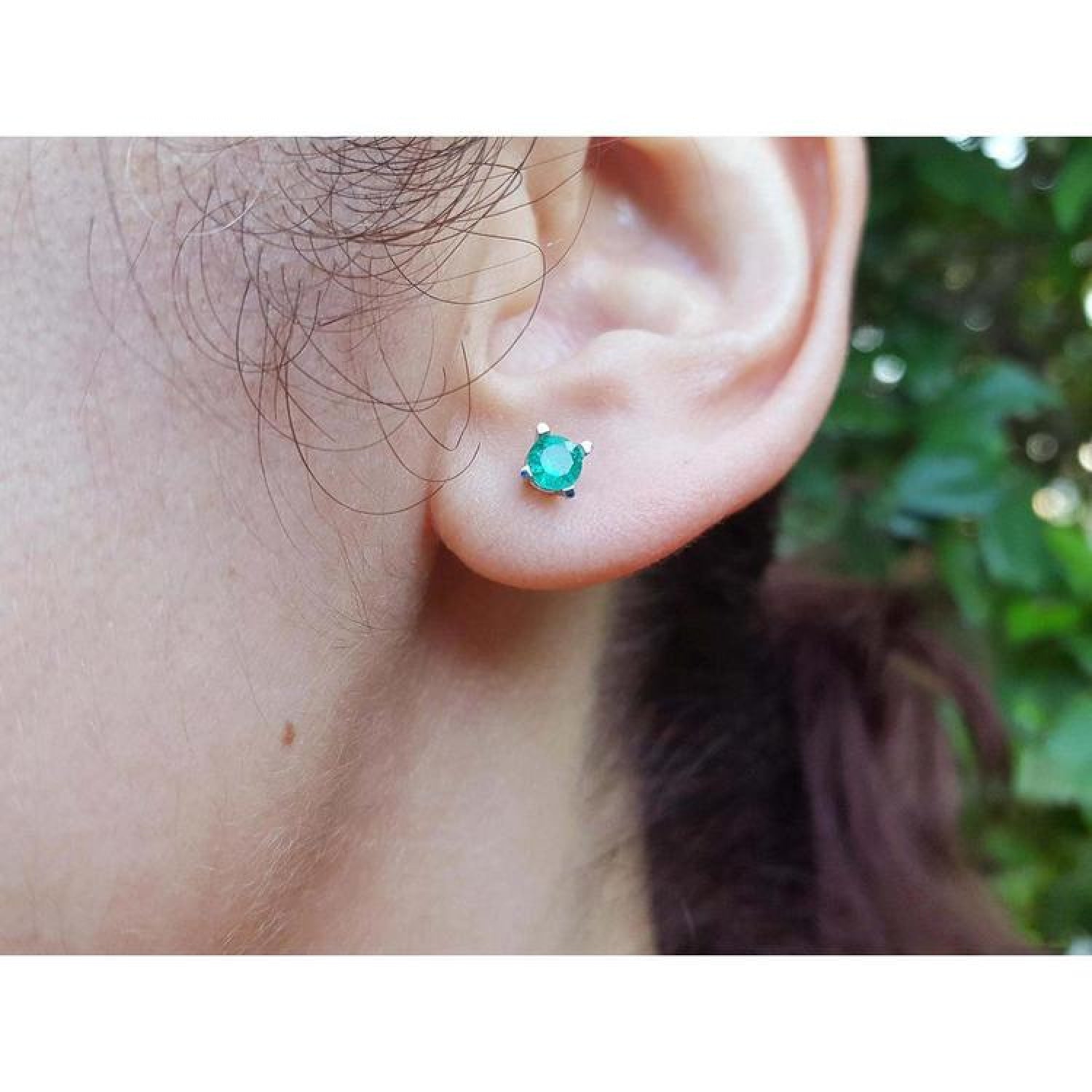 Close-up of an ear wearing a emerald stud earring with a blurred natural background.