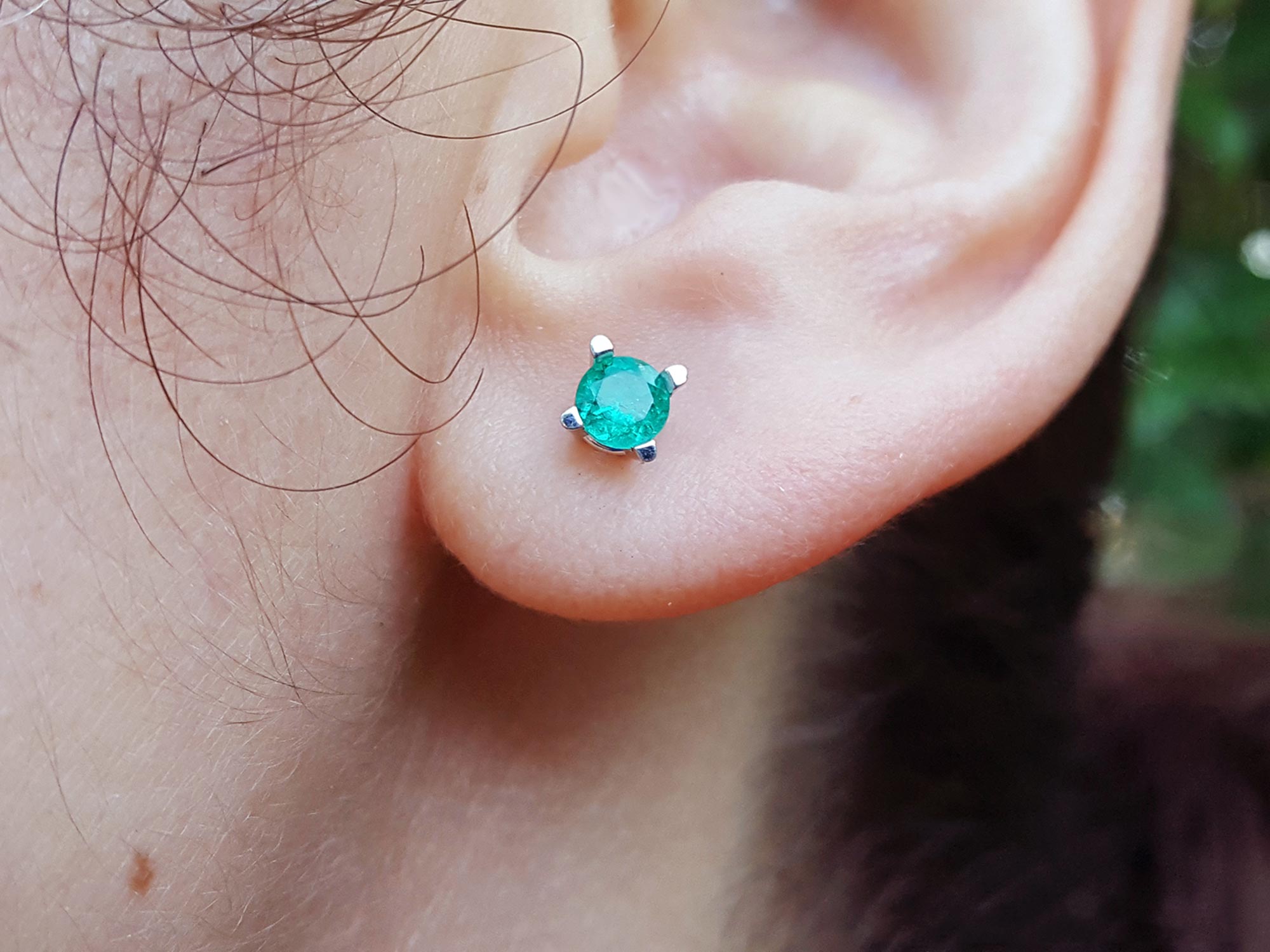 Close-up of an ear wearing a emerald gemstone earring with a blurred natural background.