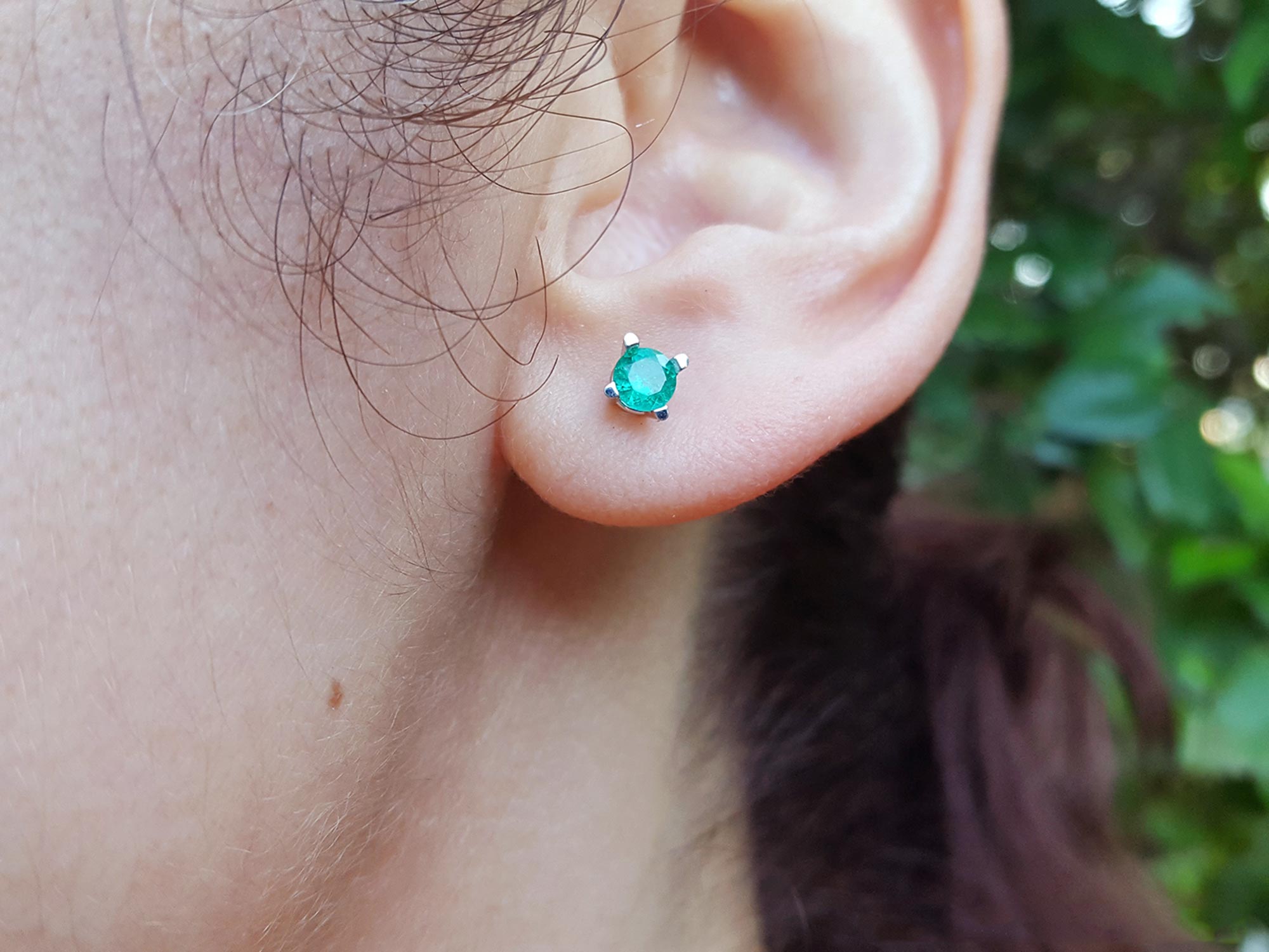 Close-up of an ear wearing a emerald stud earring with a blurred natural background.