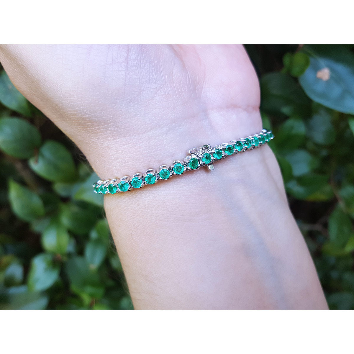 A white gold emerald tennis bracelet on a wrist with a natural background.