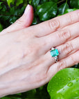 Hand wearing an emerald ring with a green leafy background.