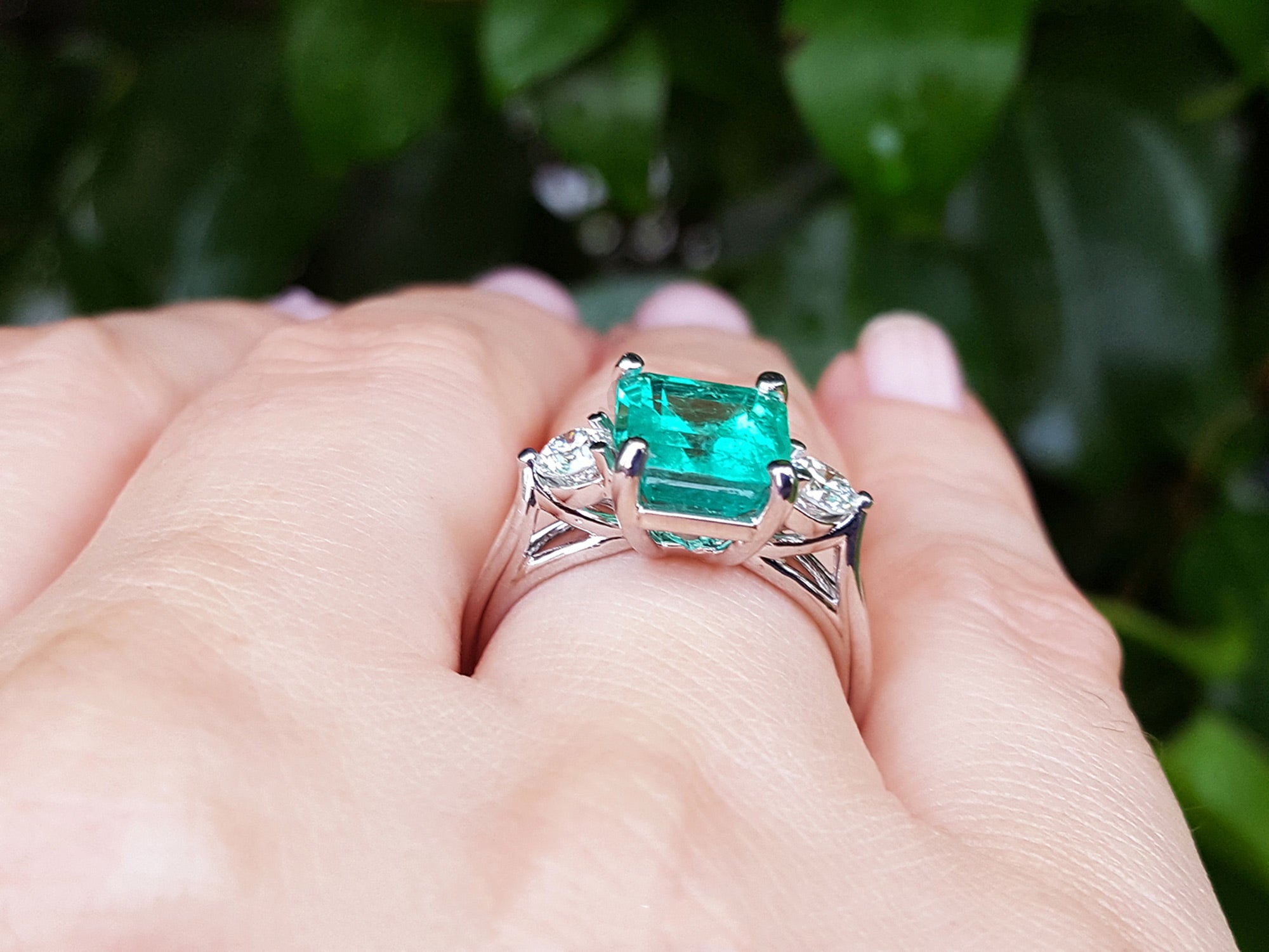 An underside view of an emerald ring with side diamonds on a hand against a blurred green leafy background.