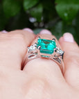 An underside view of an emerald ring with side diamonds on a hand against a blurred green leafy background.