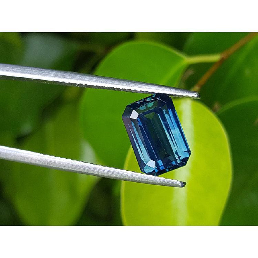 GIA certified 5.05 ct deep blue-green emerald cut sapphire held by tweezers against green leaves.