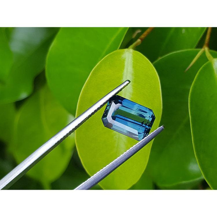 5.05 ct GIA certified deep blue-green emerald cut sapphire held by tweezers against green leaves.