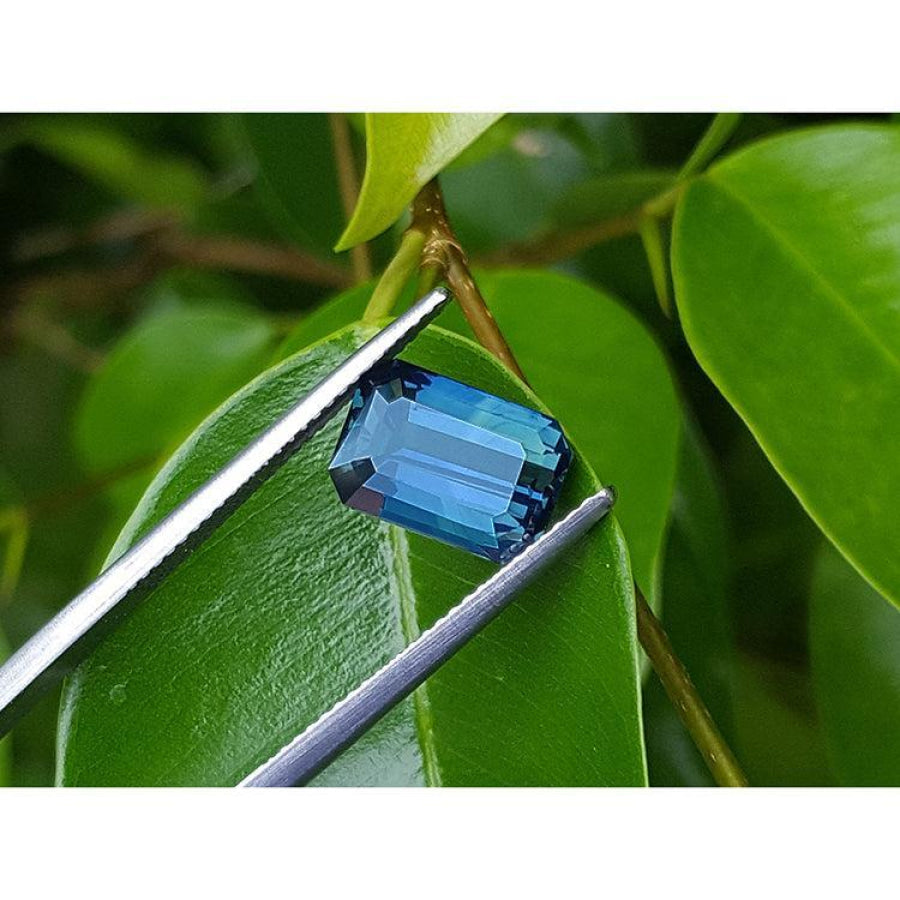 Deep blue-green emerald cut sapphire held by tweezers against green leaves.
