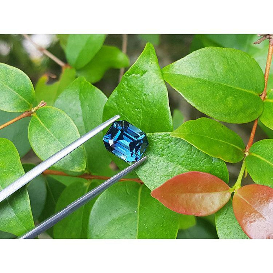 4.52 ct untreated deep blue-green emerald cut sapphire held by tweezers against green leaves.