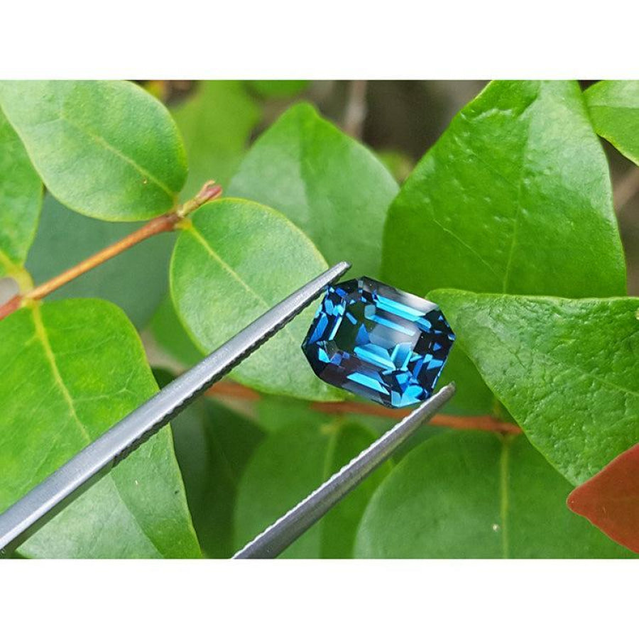 4.52 ct untreated deep blue-green sapphire, emerald cut, held by tweezers against green leaves.