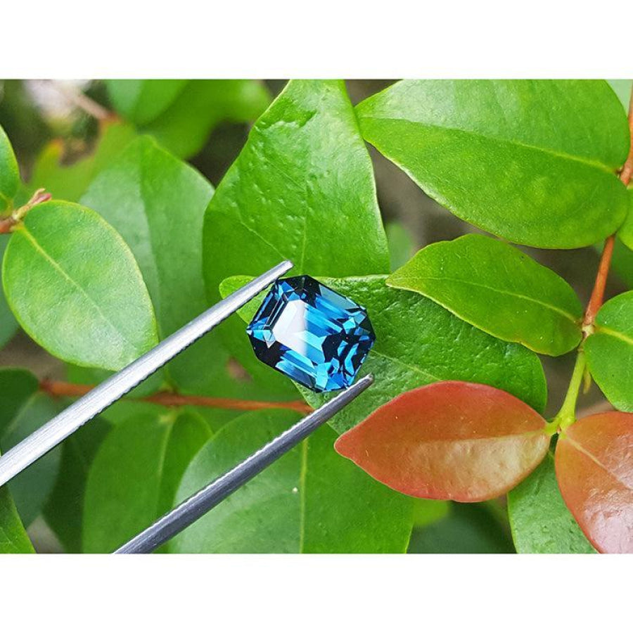 4.52 ct. untreated deep blue-green emerald cut sapphire held by tweezers against green leaves.