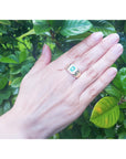 Hand wearing a yellow gold mens emerald ring against a green leafy background.