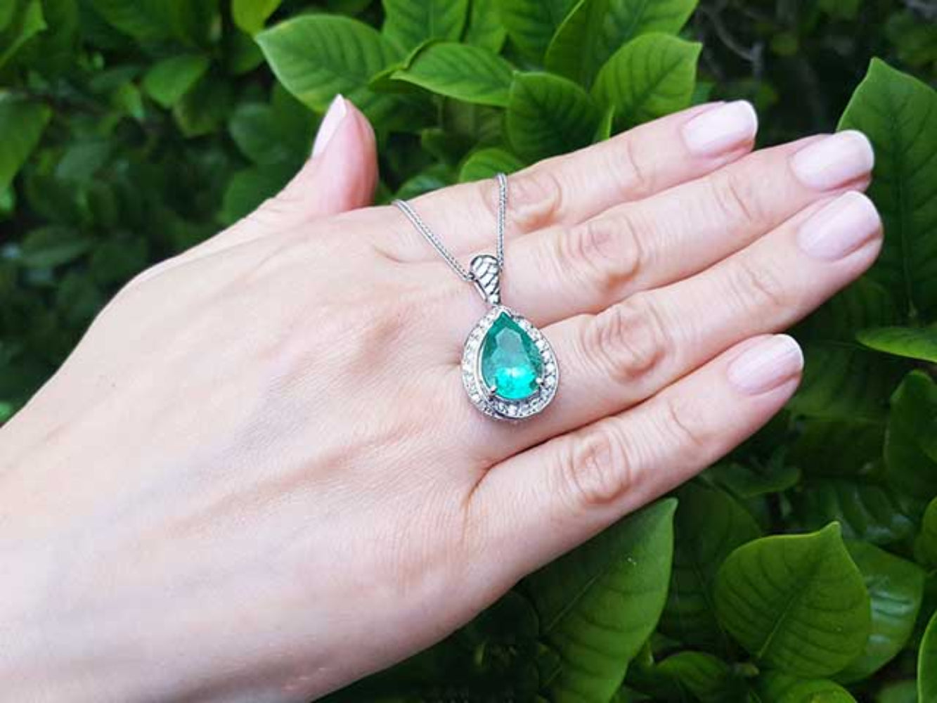 Hand holding a white gold necklace with a natural emerald pendant against a green leafy background.
