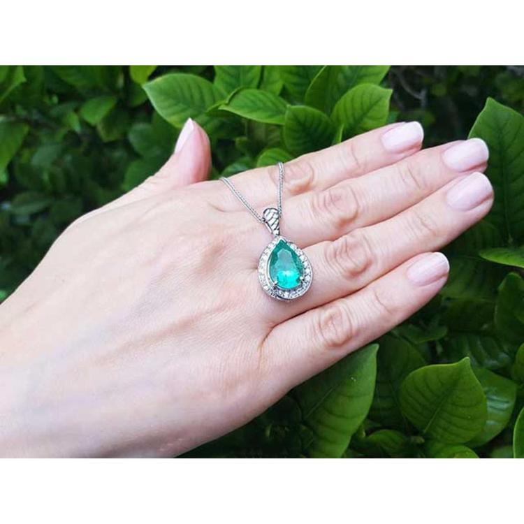 Pear-shaped natural emerald pendant with diamond halo on a 14K white gold chain, worn on a hand.